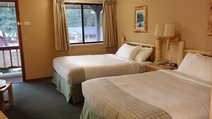 A hotel room with two beds, a wooden headboard, a window with curtains, a bedside table with a lamp, and a small table with a chair. - Wildwood Inn (Twain Harte)
