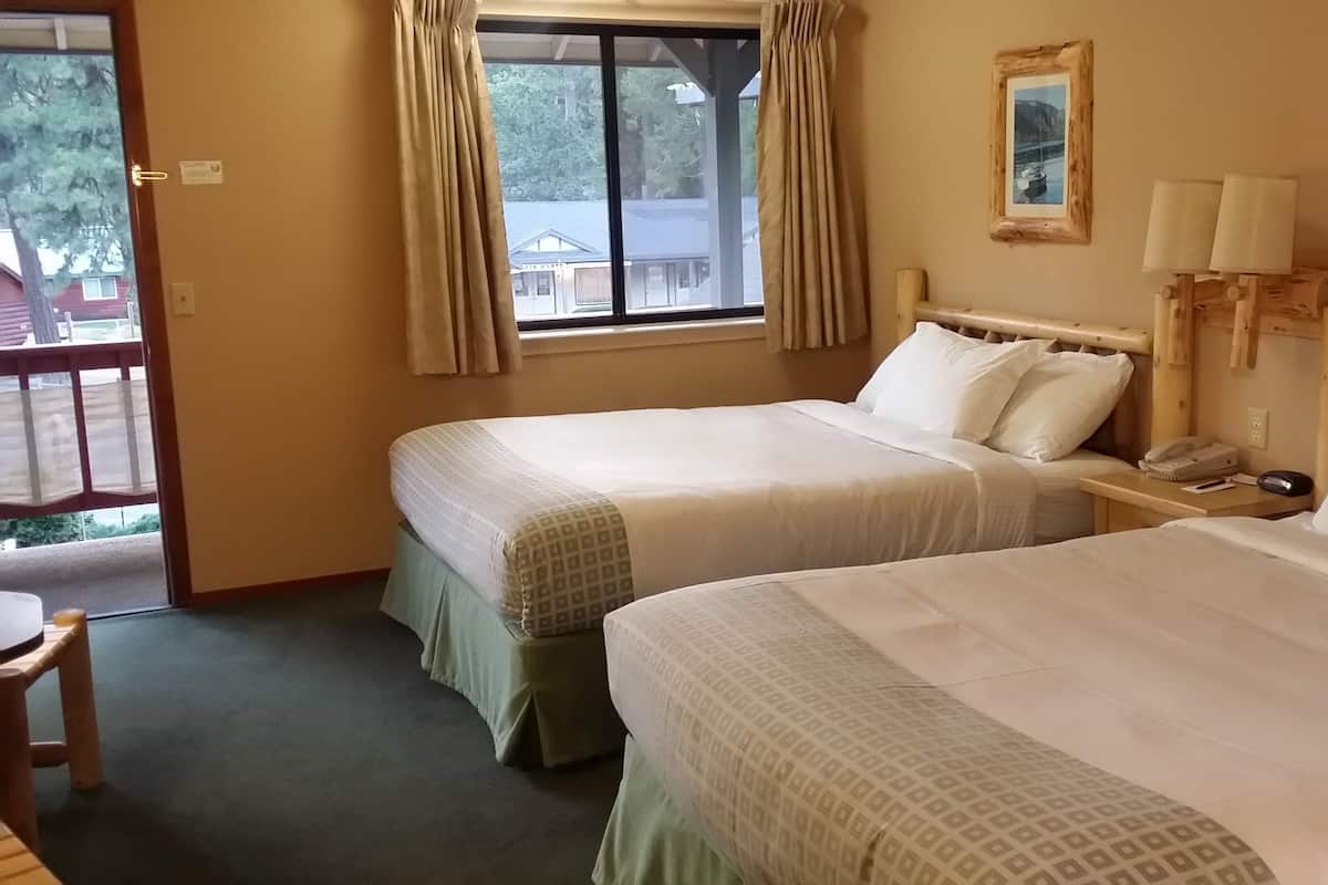 A hotel room with two beds, a wooden headboard, a window with curtains, a bedside table with a lamp, and a small table with a chair.