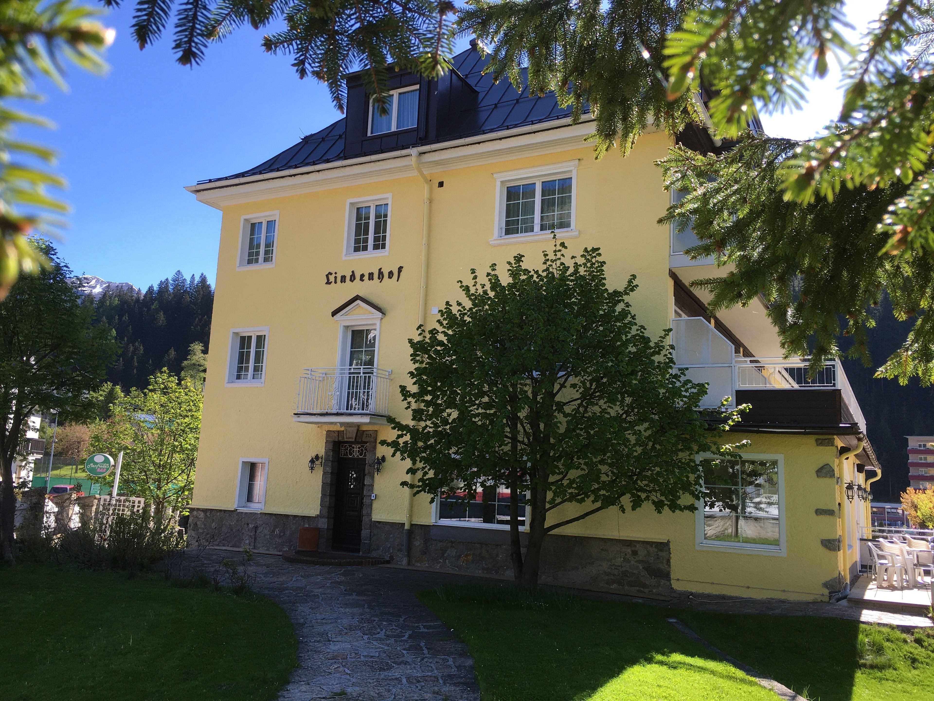 Un bâtiment jaune dont la façade porte l’inscription « Lindenhof », avec une entrée en pierre et un balcon agrémenté de balustrades.