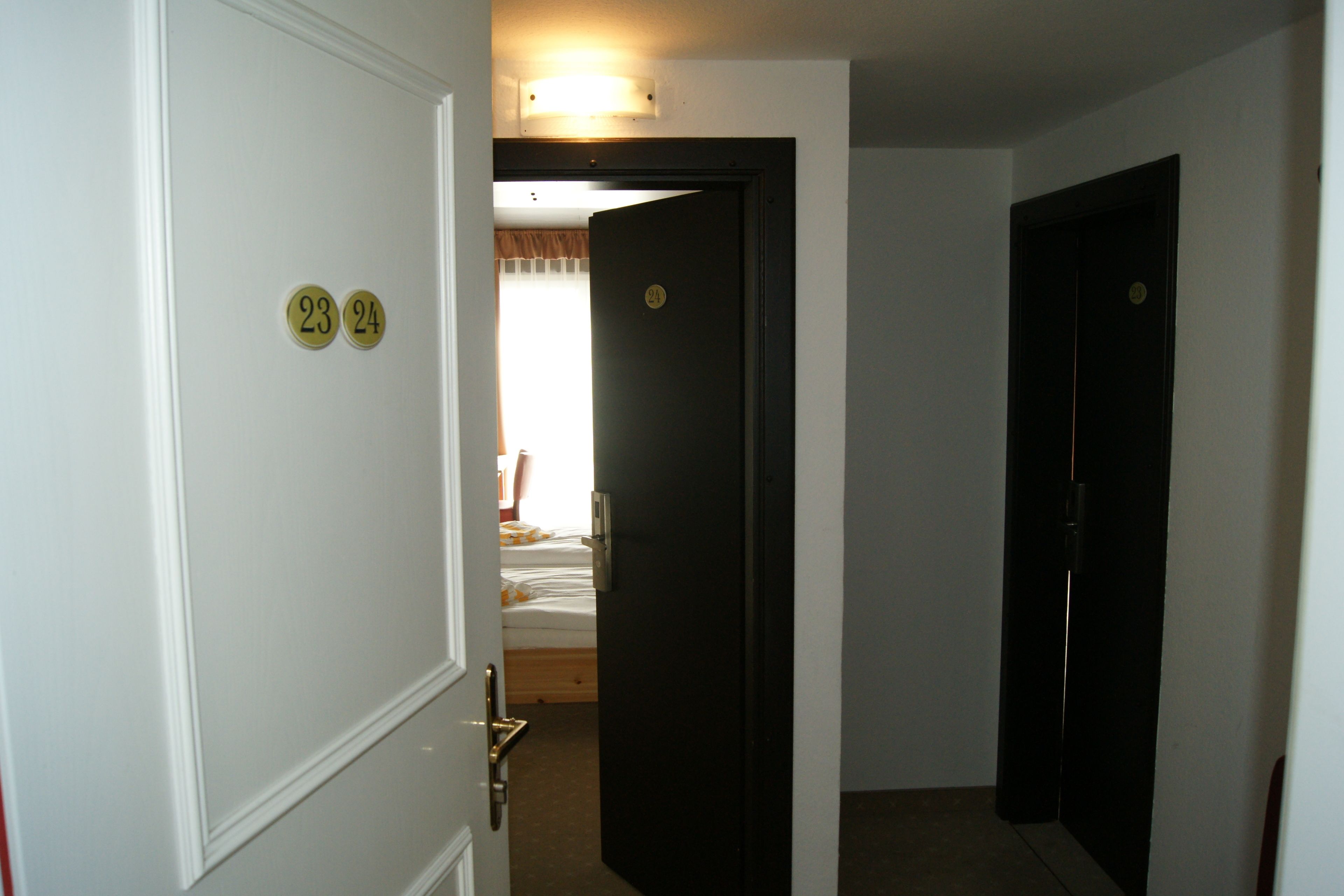 A narrow hallway with two doors, one marked with numbers 23 and 24, leading to a bedroom with a bed and curtains.