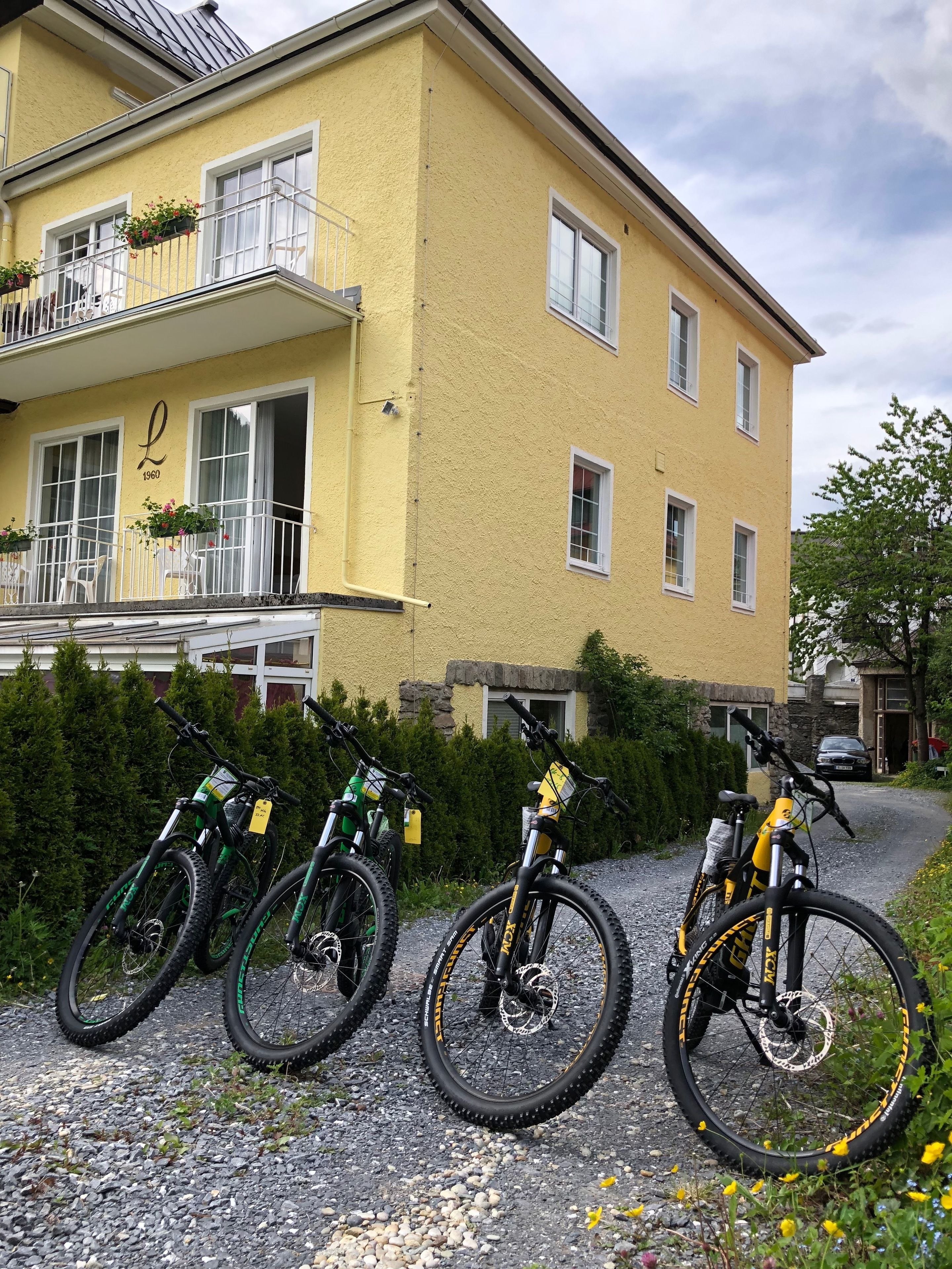 A yellow building with a balcony, bicycles parked in front, and a gravel path.