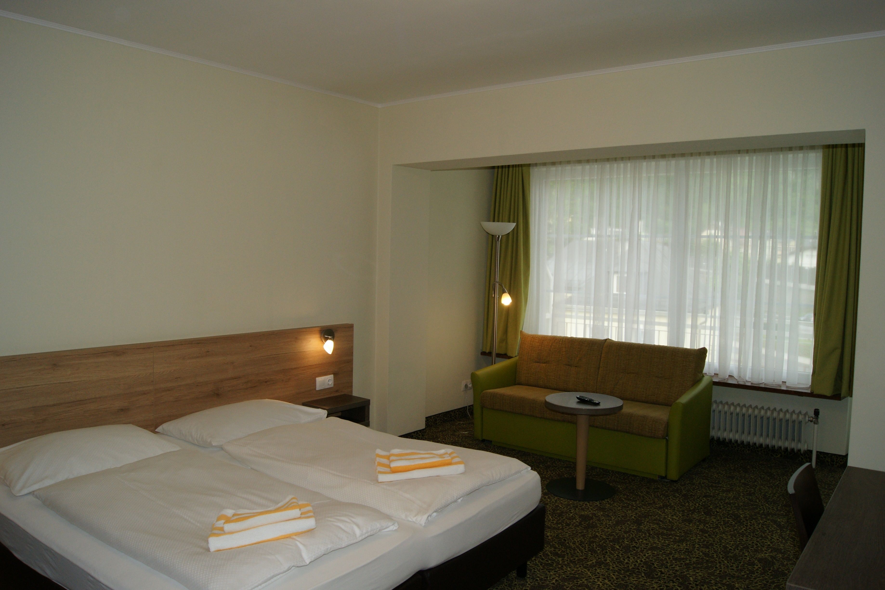 Habitación de hotel con una cama doble, un sillón verde, una mesita y una ventana con cortinas traslúcidas.