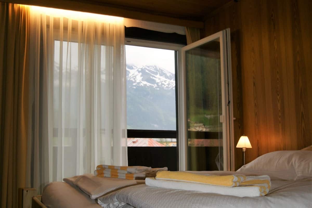 A wooden-framed bed with white bedding, a window with curtains, and a view of snow-capped mountains.