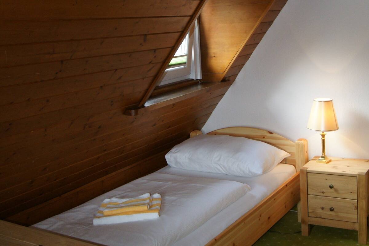 A small, cozy bedroom with a wooden bed, a bedside table with a lamp, and a window.