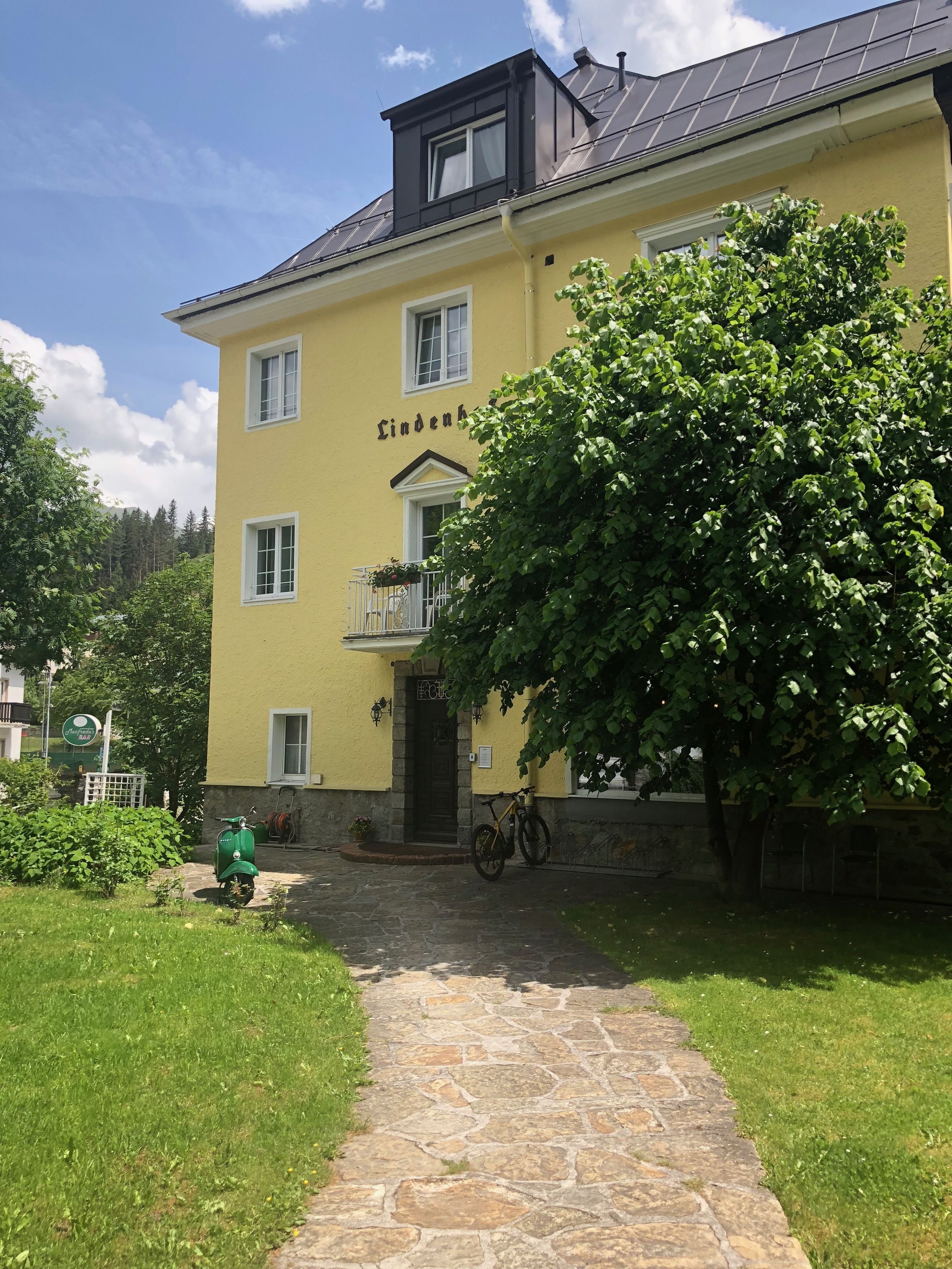 Un edificio amarillo con el nombre 'Lindenhof' en la fachada, un sendero de piedra que conduce a la entrada y una bicicleta estacionada afuera.