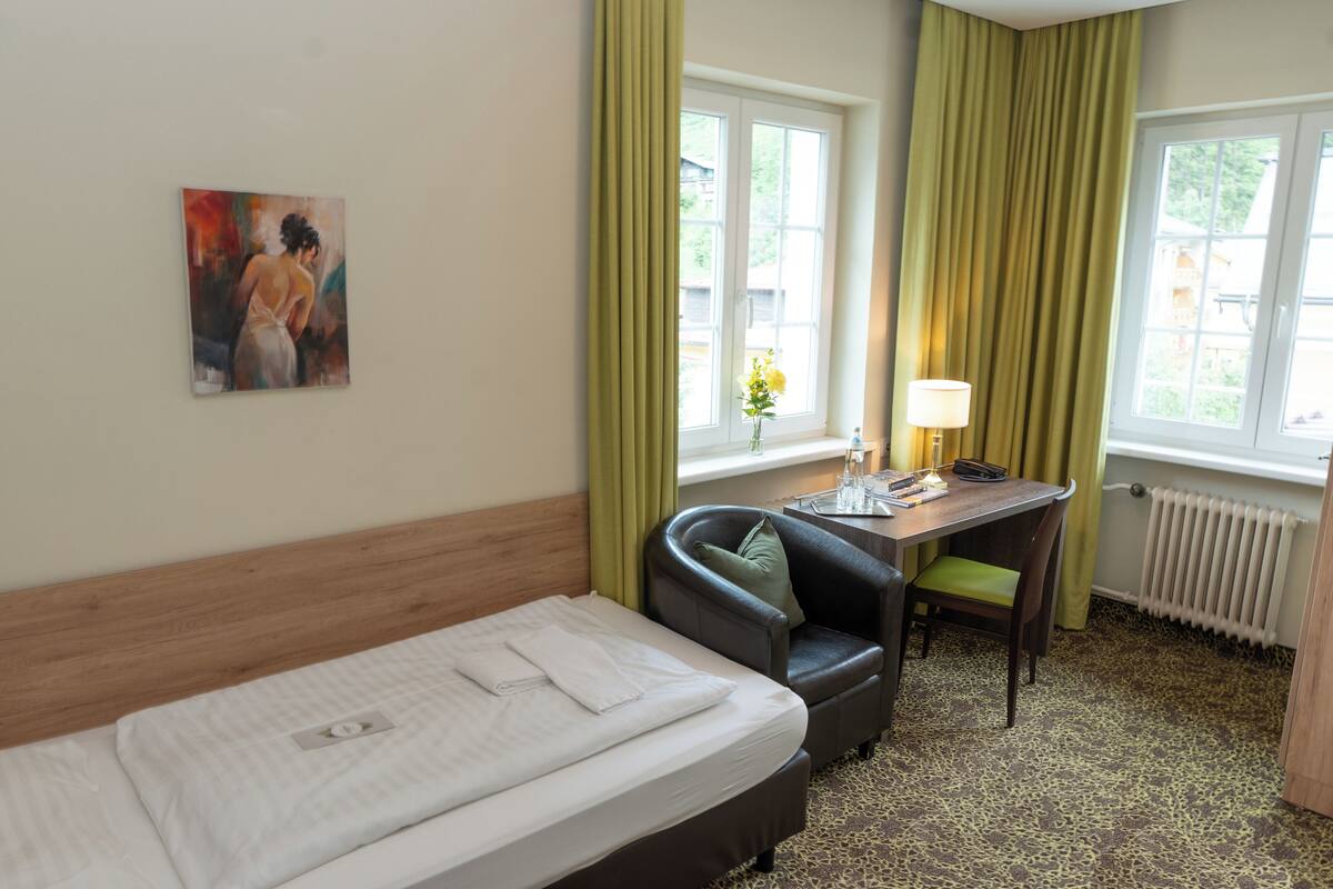 A hotel room with a bed, a desk, a chair, a painting on the wall, and a window with curtains.