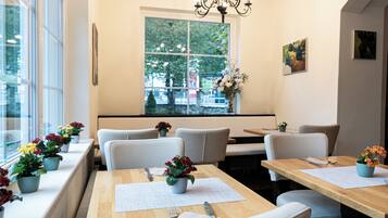 A restaurant with a long counter, multiple tables, and chairs. There are potted plants on the windowsill and a chandelier above the counter.