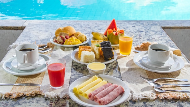 A breakfast table with various foods, drinks, and utensils by a poolside. - Garça Branca Praia Hotel (Porto Seguro)