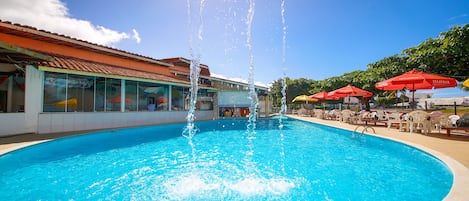Piscina com jatos de água, rodeada por espreguiçadeiras e guarda-sóis, e um edifício com telhado de azulejos ao fundo.