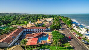 An aerial view of a coastal resort with a swimming pool, beachside villas, and a road leading to the beach. - Garça Branca Praia Hotel (Porto Seguro)