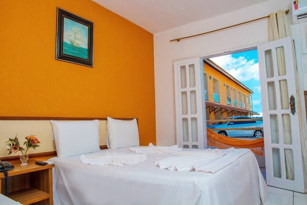 A hotel room with a bed, a nightstand, a framed picture on the wall, and a view of a street with buildings and a car.