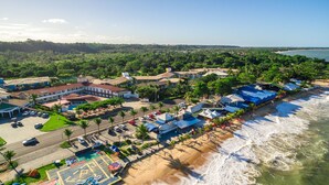 An aerial view of a coastal town with a beachfront, buildings with blue roofs, and a road with parked cars. - Garça Branca Praia Hotel (Porto Seguro)