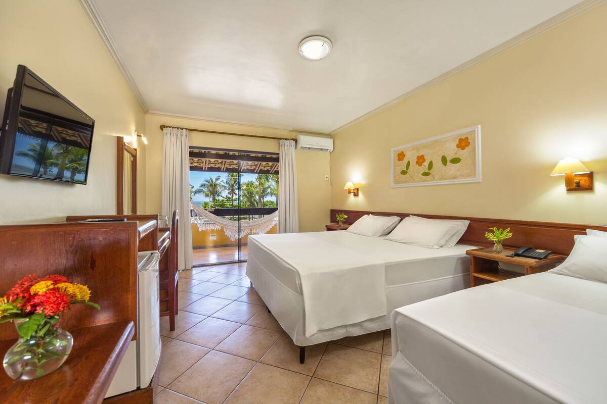 A hotel room with two beds, a desk with a vase of flowers, a view of palm trees through a window, and a hammock.