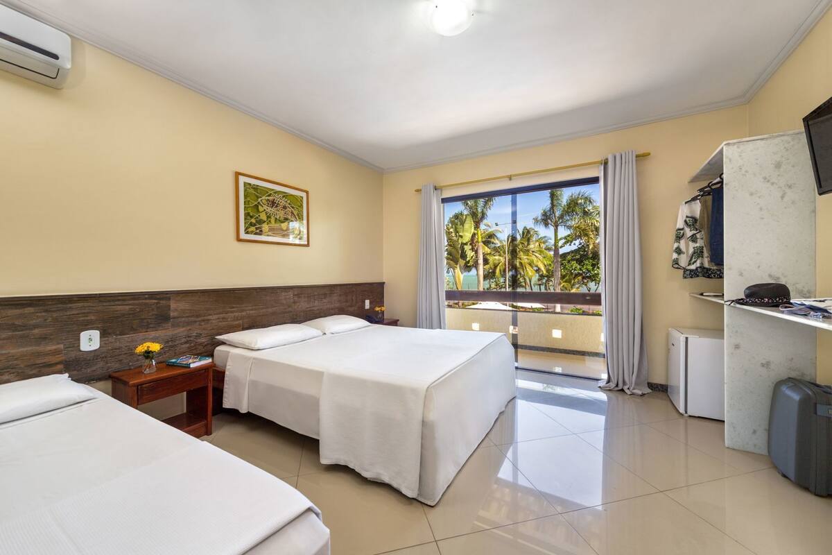 A hotel room with two beds, a TV, a minibar, and a balcony with a view of palm trees.
