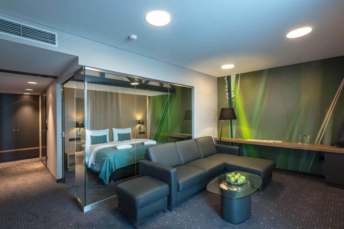 A modern hotel room with a glass partition separating the sleeping area from the living space, a sofa, a small table, and a green wall with a plant.