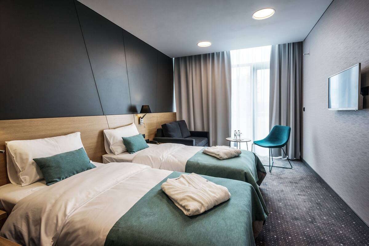 A hotel room with two beds, a small table, a chair, a TV mounted on the wall, and a large window with curtains.