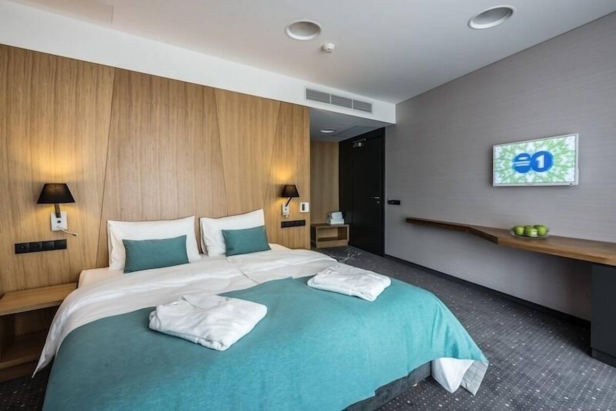 A modern hotel room with a large bed, two bedside tables, a wall-mounted TV, and a small shelf with fruits.