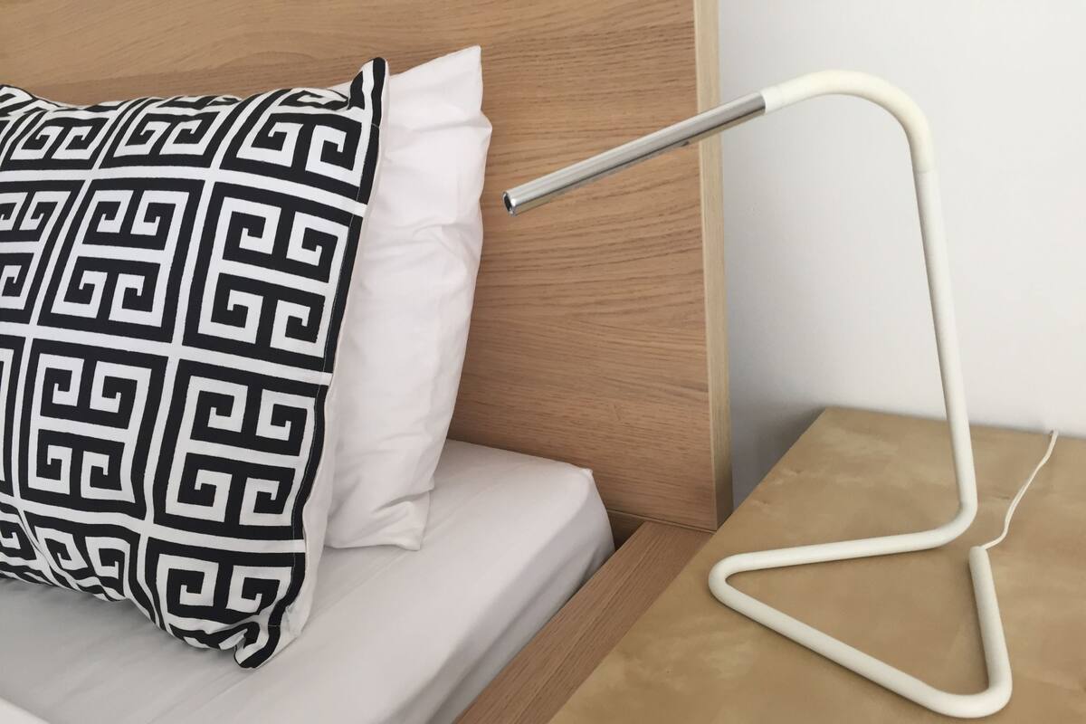 A neatly made bed with a white mattress and a black and white patterned pillow. A white bedside lamp with a metallic base is on a wooden nightstand beside the bed.