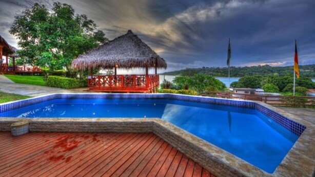 Piscina con un pabellón de techo de paja, rodeada de exuberante vegetación y vistas a una masa de agua.