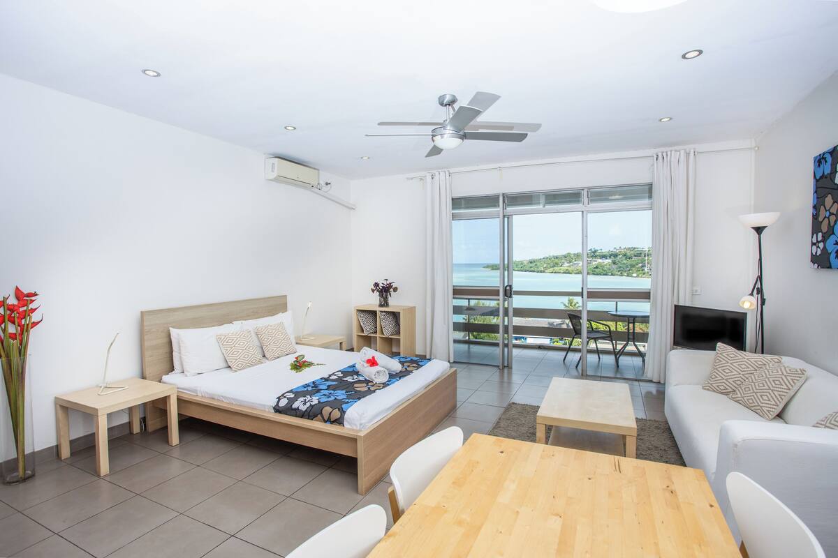 A modern hotel room with a bed, a wooden table, a sofa, and a balcony with a view.