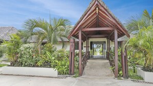 Une allée couverte mène à un bâtiment dont le toit est fait de chaume, et qui est entouré d’une végétation tropicale luxuriante.