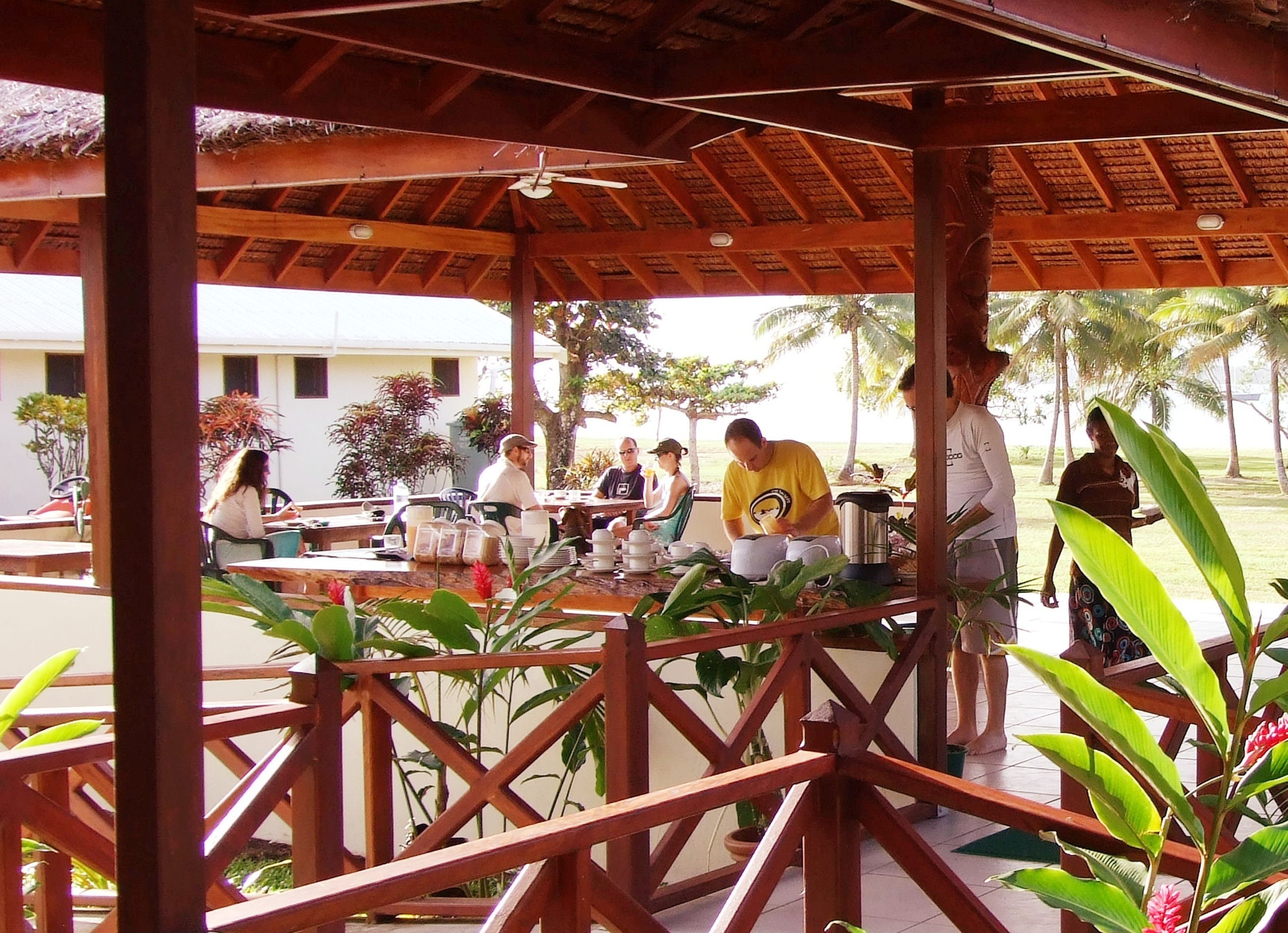A covered outdoor dining area with wooden beams and a thatched roof, surrounded by lush greenery and palm trees.