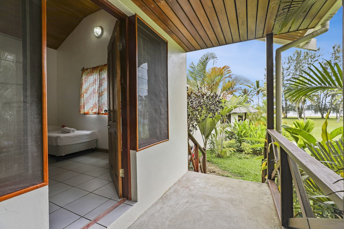 A room with a bed, a window with a curtain, and a view of a garden with palm trees.