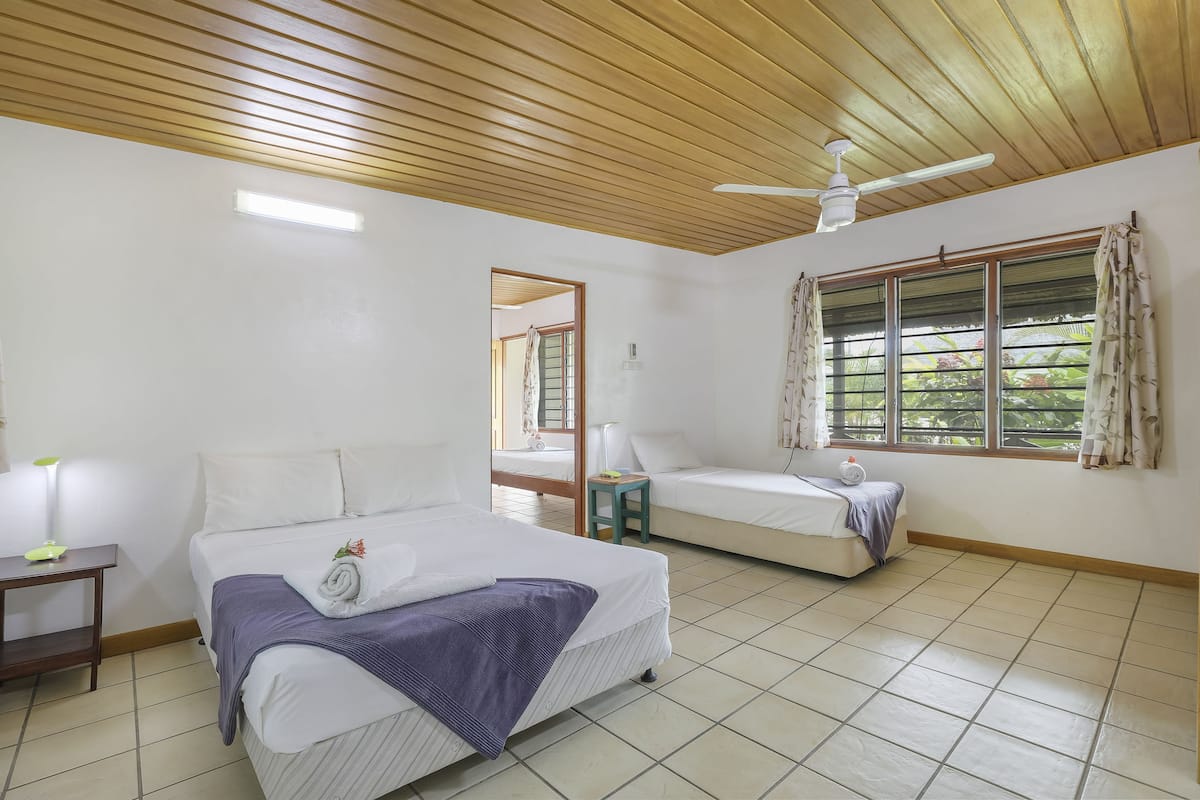 A bedroom with two beds, a ceiling fan, a window with curtains, and a nightstand with a lamp.