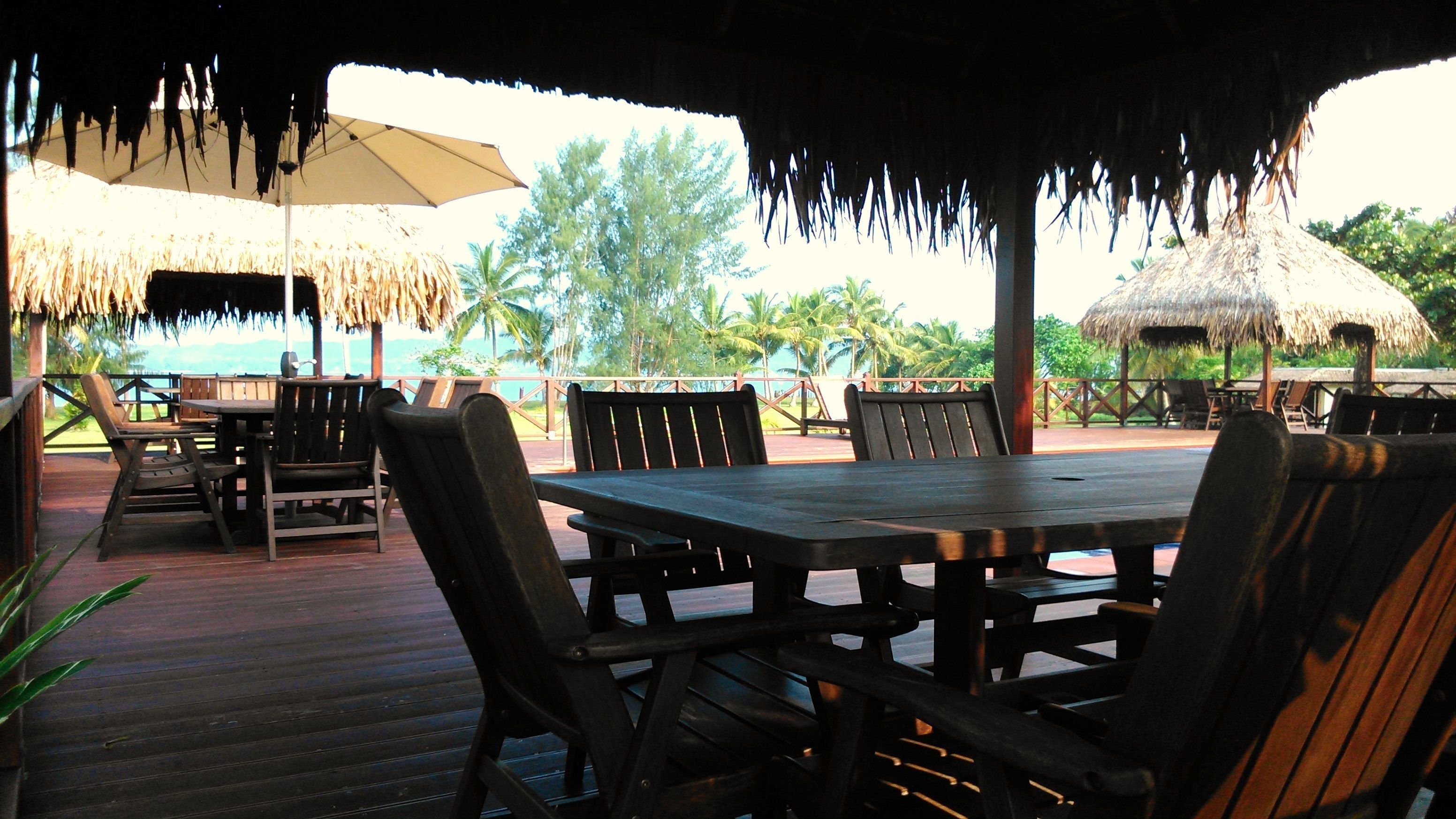Un área de comedor al aire libre con mesas y sillas de madera, sombrillas de paja y vistas a palmeras y al océano.