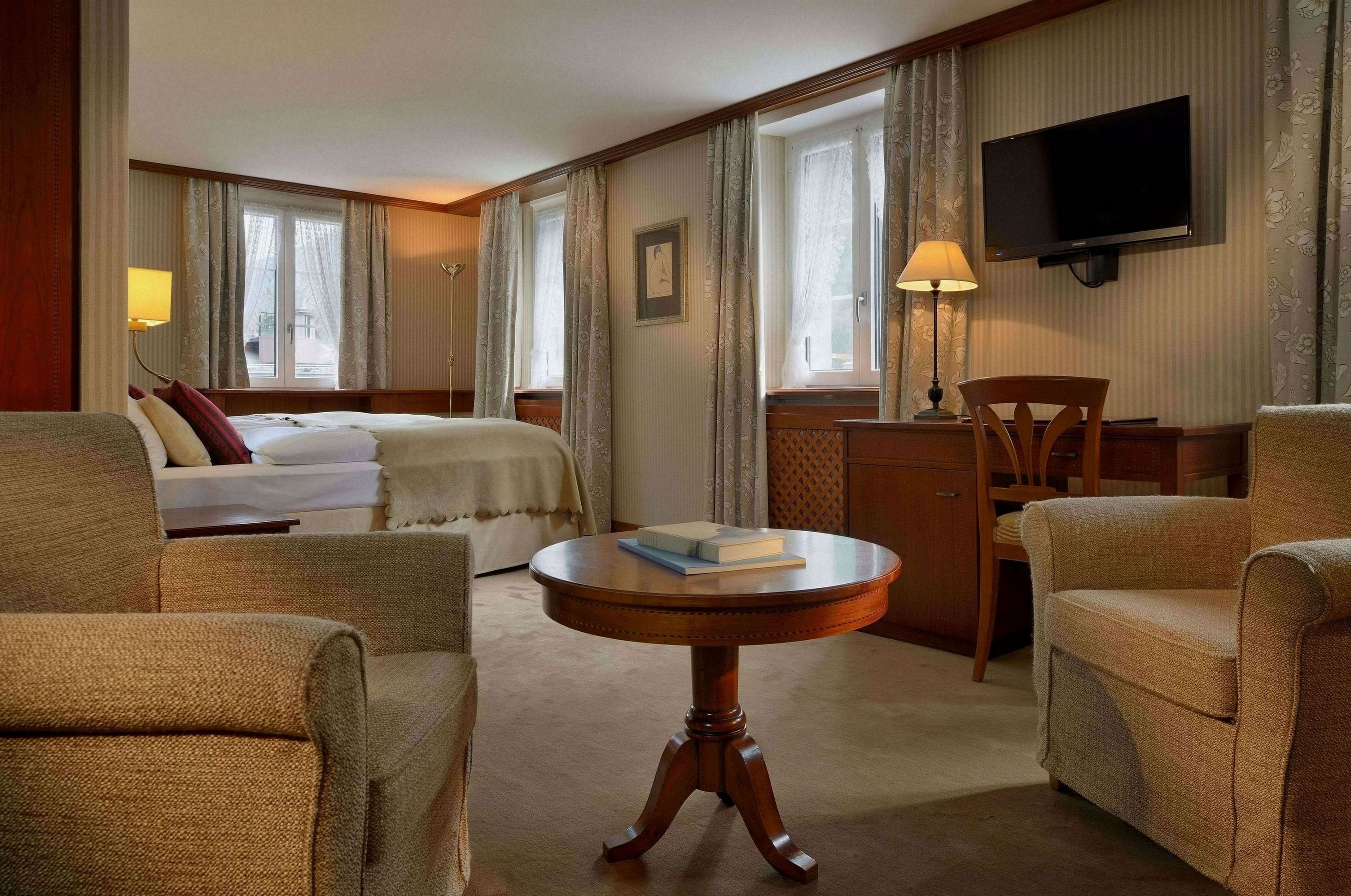 A hotel room with a bed, two armchairs, a small round table with a book, a TV mounted on the wall, and a desk with a lamp.
