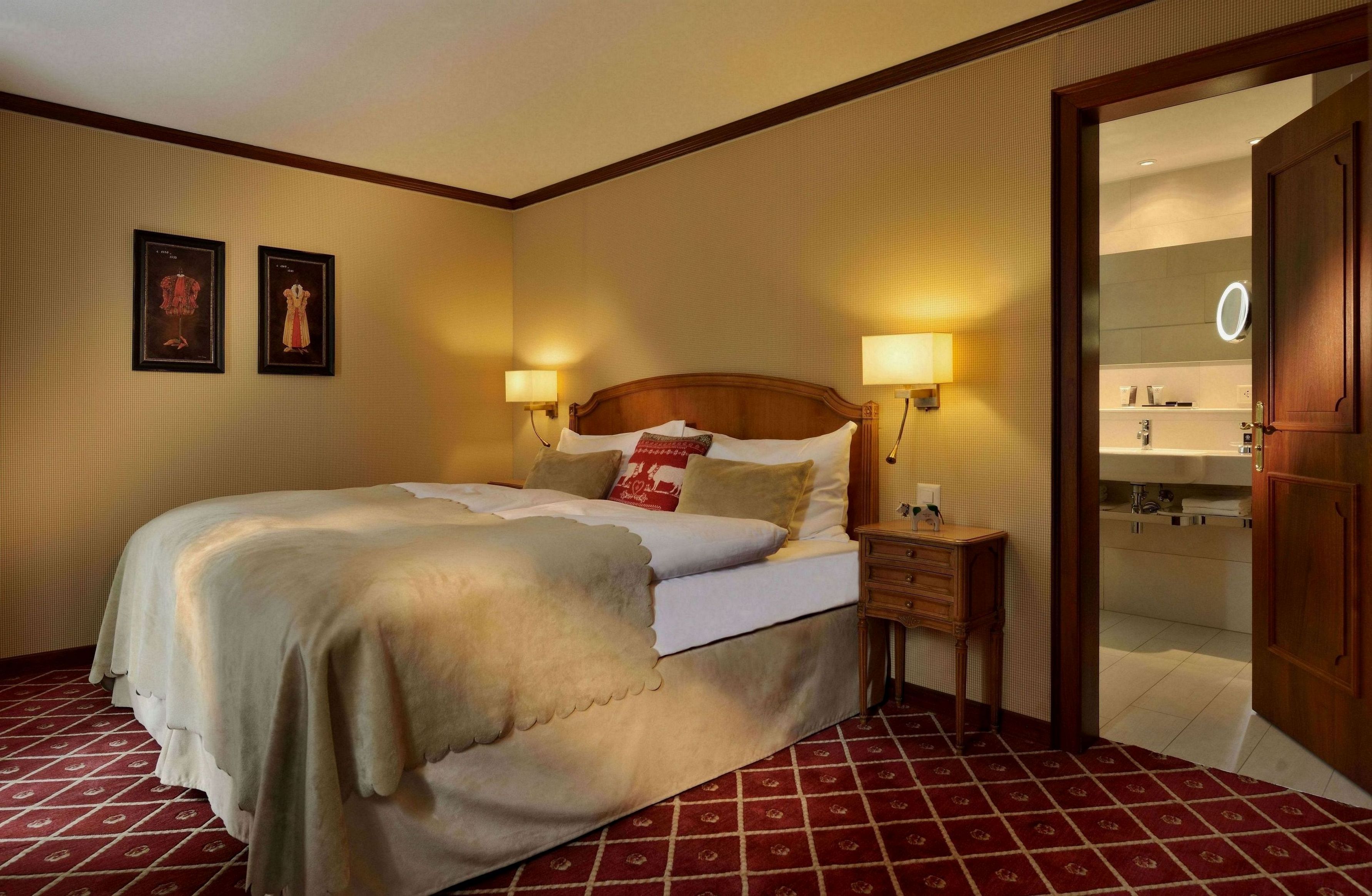A hotel room with a large bed, two bedside lamps, a nightstand, and a door leading to a bathroom.