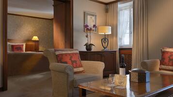 A hotel room with a bed, two armchairs, a table with wine glasses, a lamp, and a potted plant.