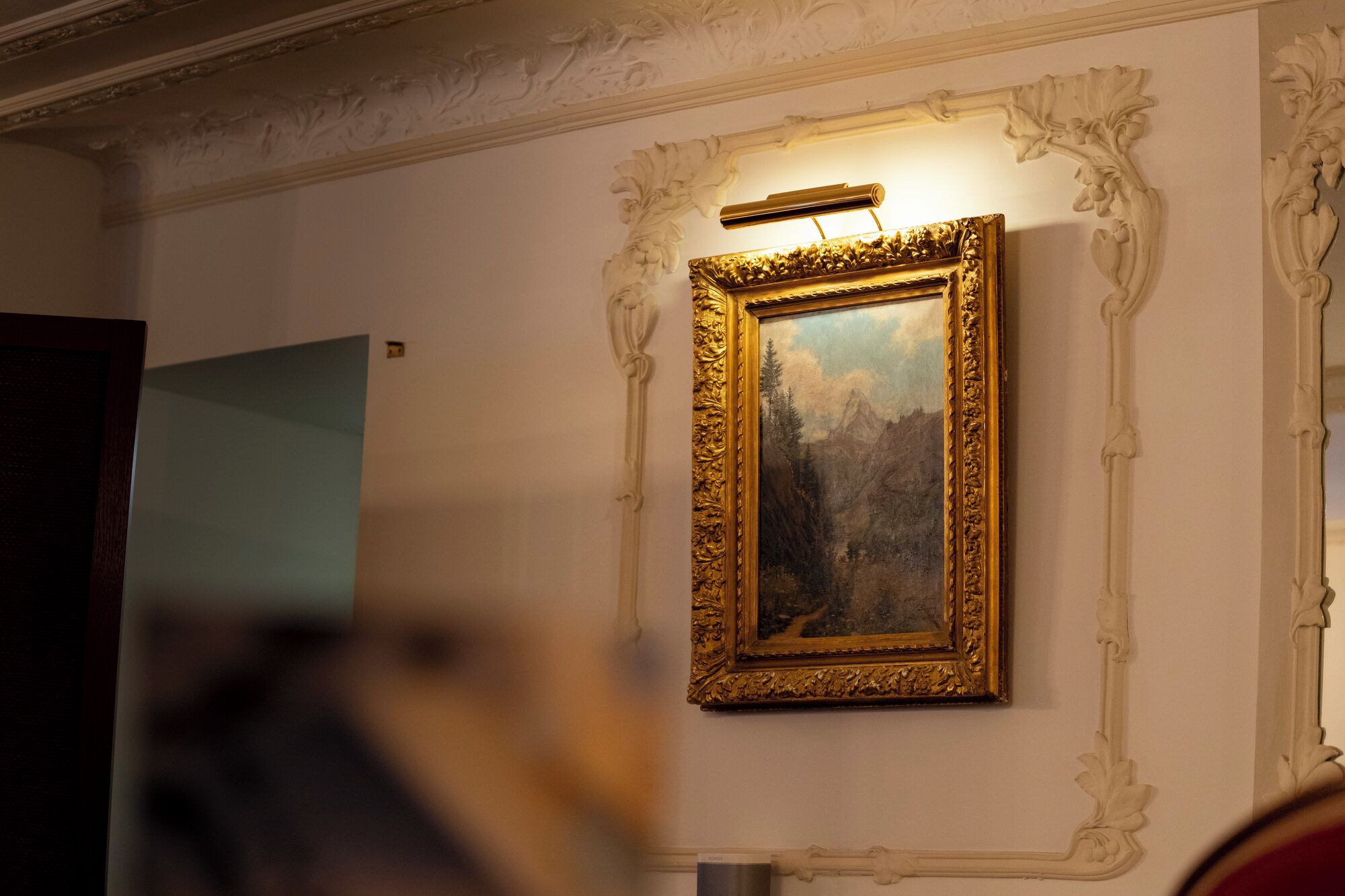 A framed painting with a gold frame, mounted on a wall with ornate molding.