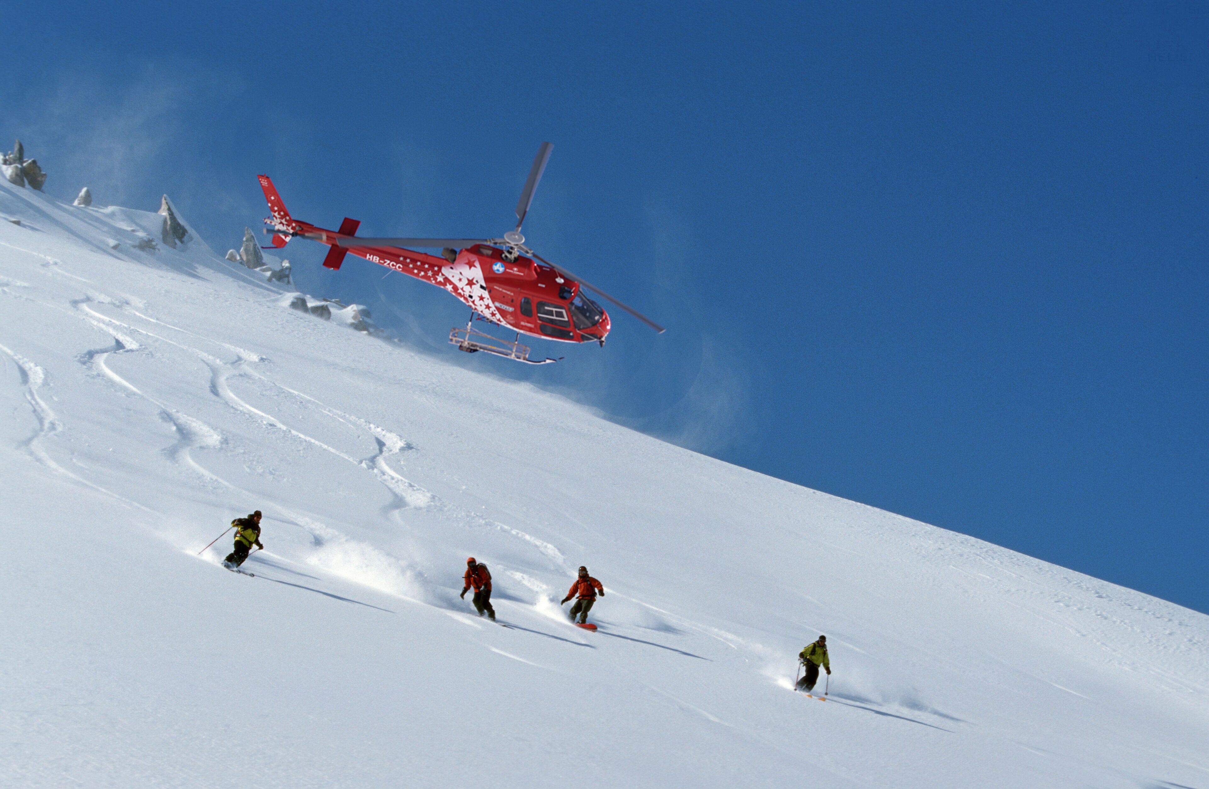 A red helicopter flying over a snowy mountain slope with skiers below.