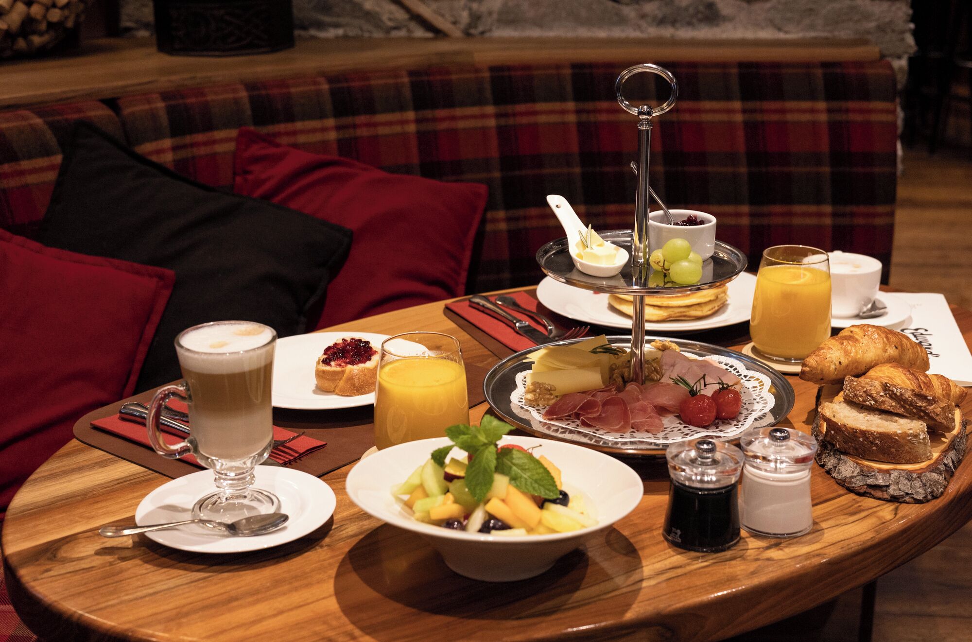 A breakfast buffet with various dishes, including fruits, pastries, and a coffee drink, set on a wooden table in a cozy room with a plaid sofa.