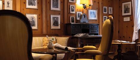A cozy room with wooden paneling, a round table, two chairs, a lamp, and framed pictures on the walls.