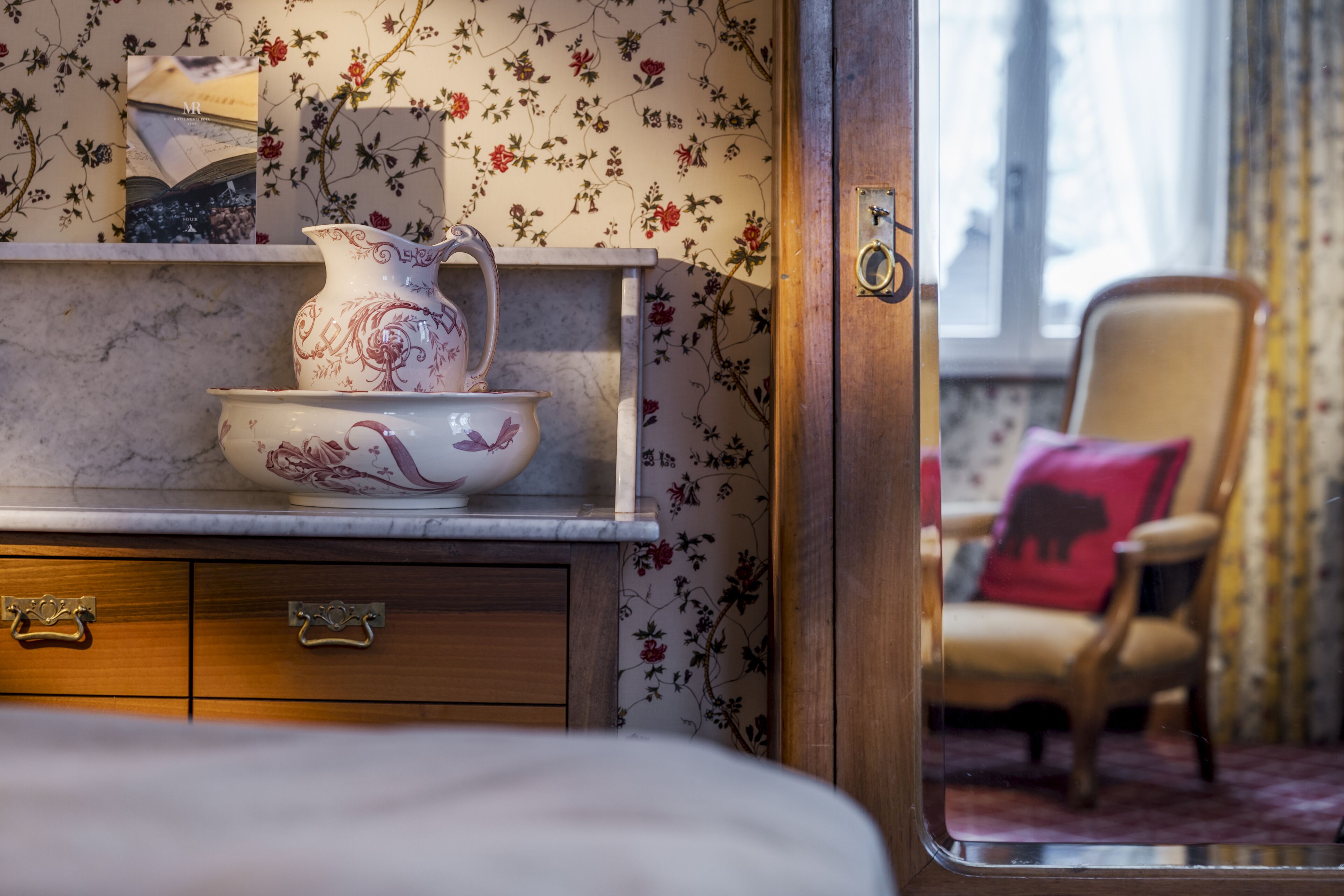 Una habitación con encimera de mármol, un mueble de madera con cajones, papel tapiz floral, un cojín rojo en una silla y una ventana con cortinas.