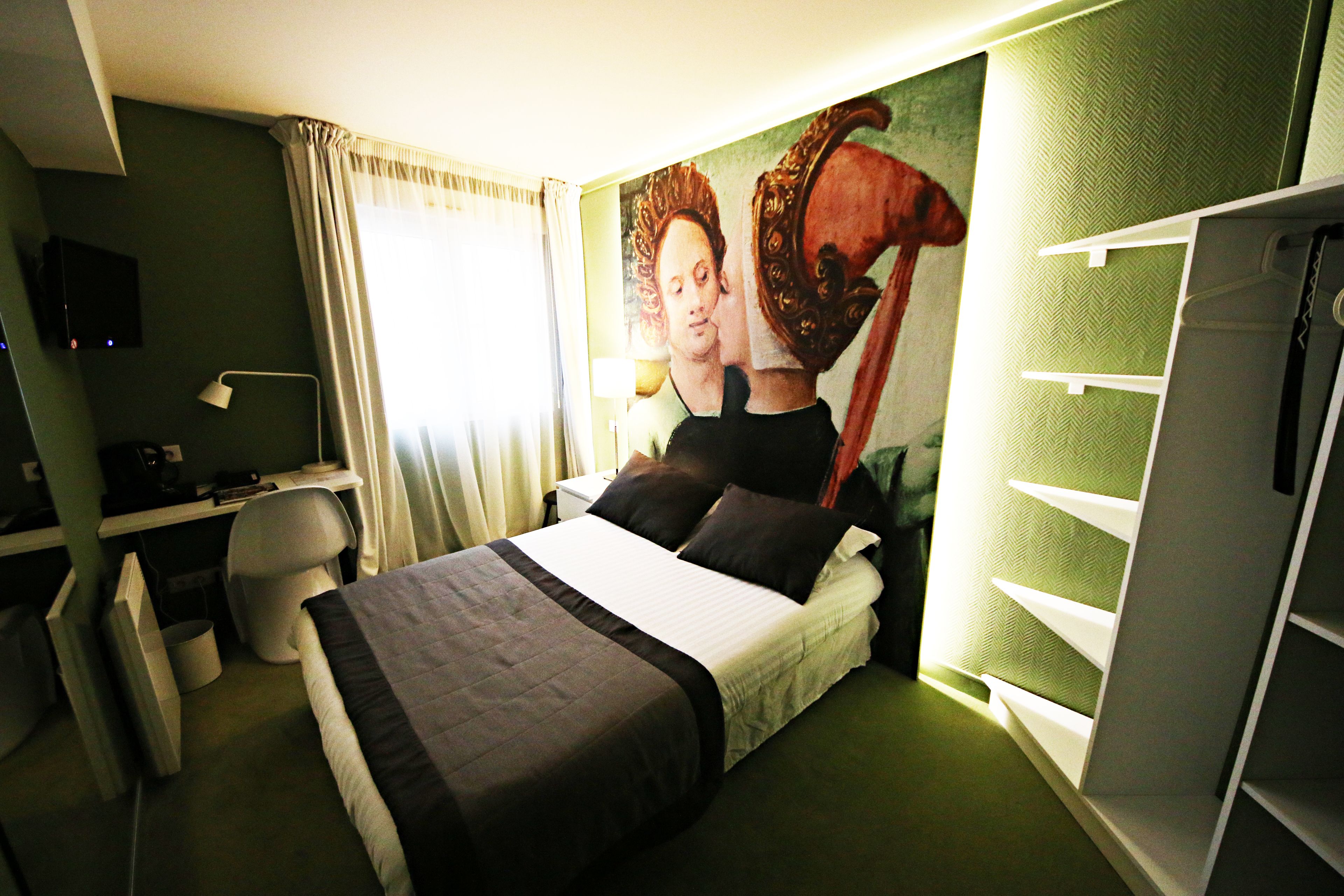 A hotel room with a bed, a desk with a computer, a chair, a TV, and a painting on the wall.