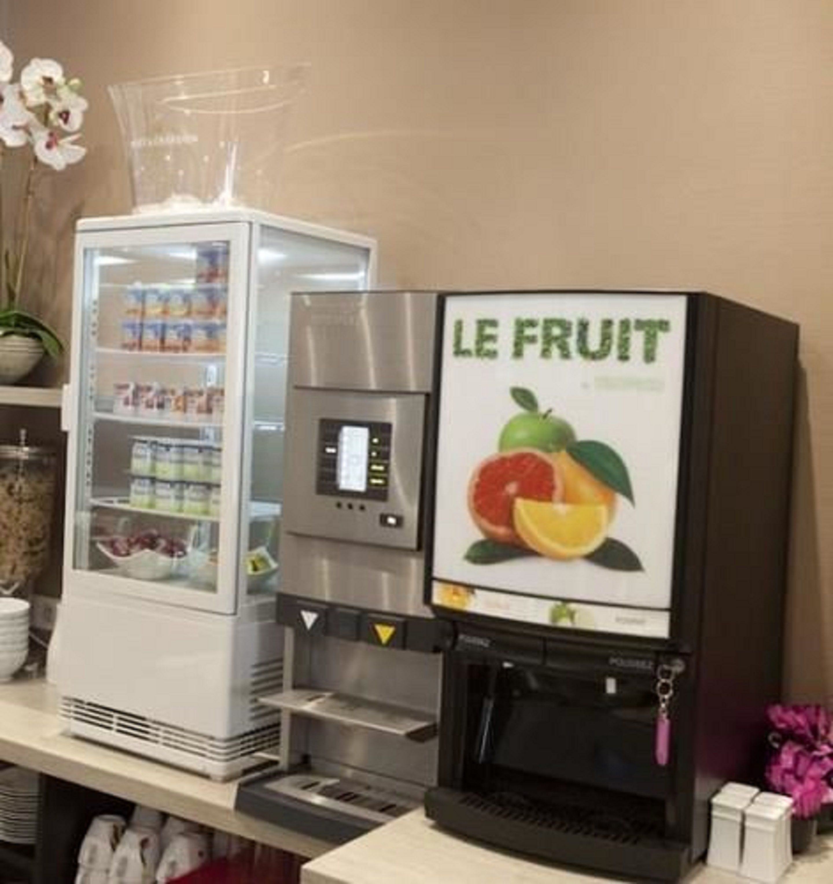 A self-service beverage dispenser with a fruit-themed sign, a refrigerated beverage cooler, and a potted orchid.