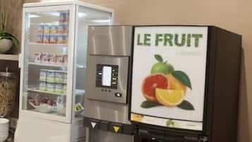 A self-service beverage dispenser with a fruit-themed sign, a refrigerated beverage cooler, and a potted orchid.