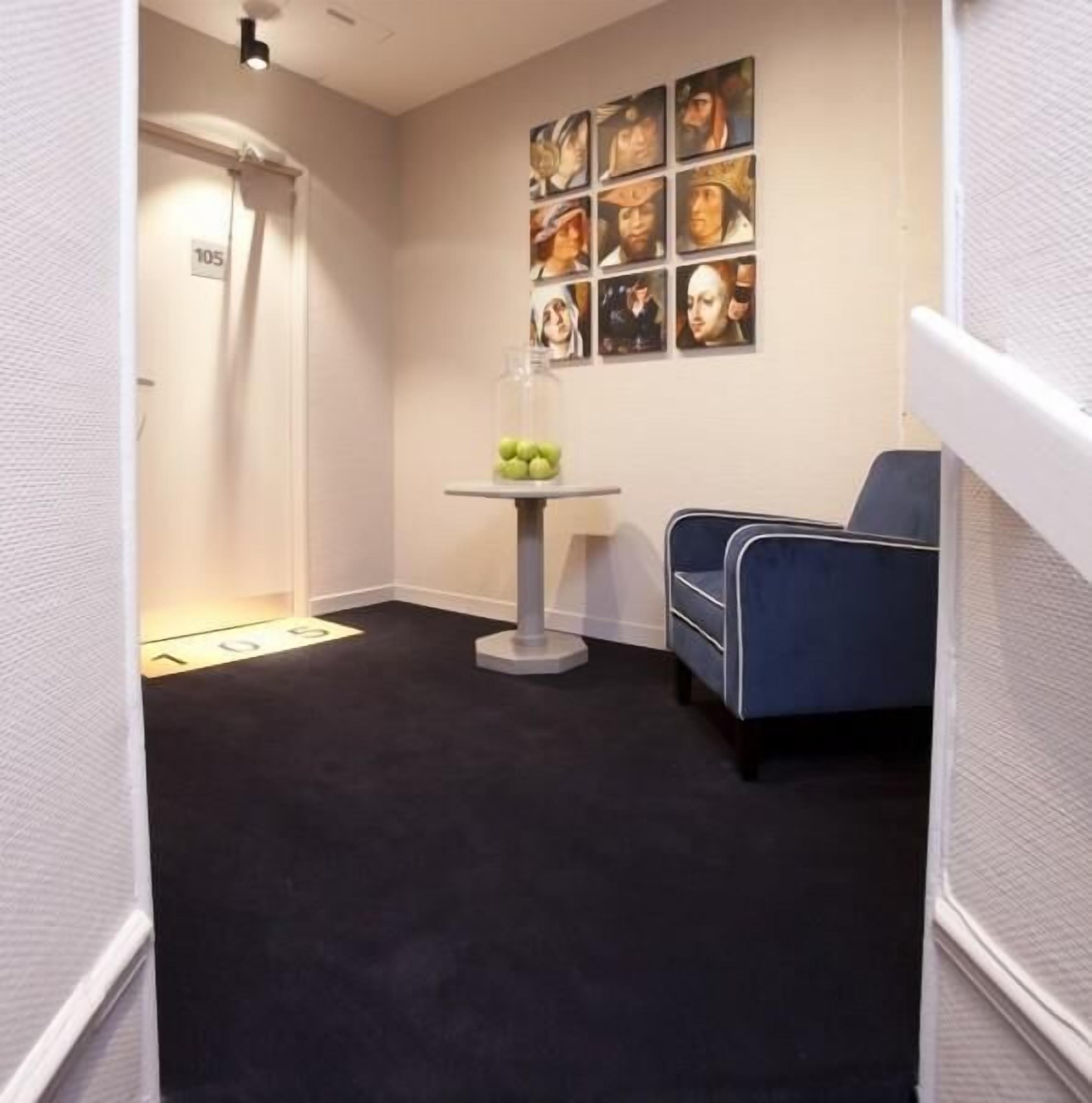 A hotel room with a black carpet, a blue chair, a small table with a glass top, and a wall with multiple framed pictures.
