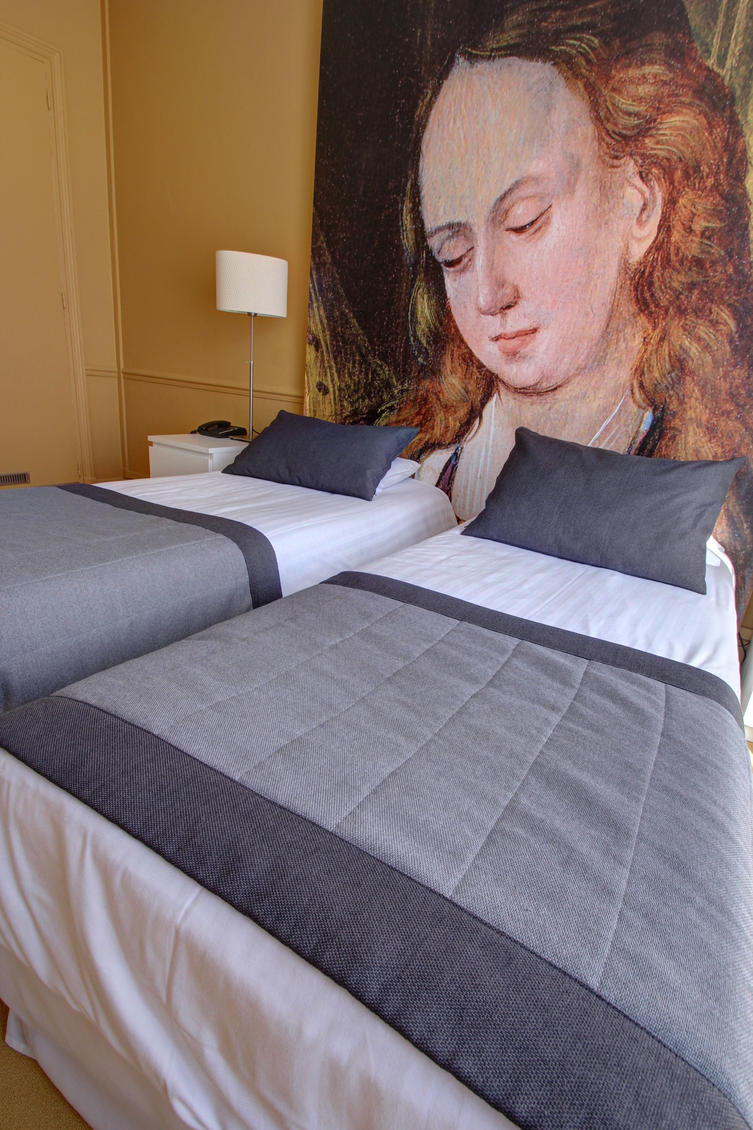 Un lit double avec une literie aux tons de gris et de blanc, une table de chevet avec une lampe, et un grand tableau accroché au mur.