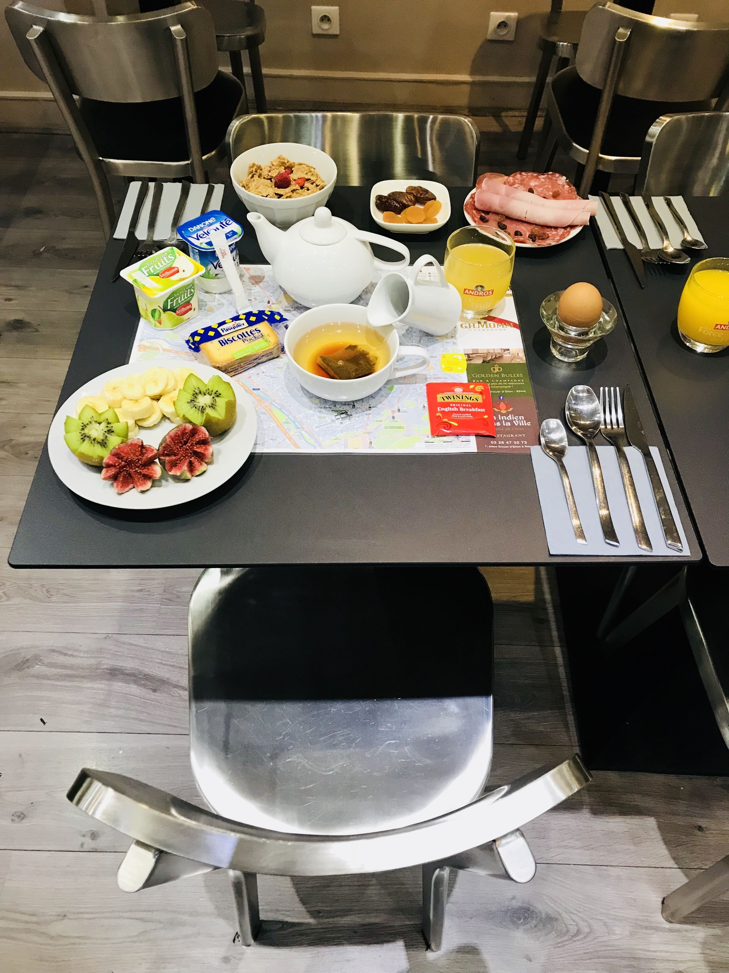 Uma mesa de café da manhã com diversos alimentos, incluindo frutas, uma tigela de cereal, um bule de chá e um copo de suco de laranja.