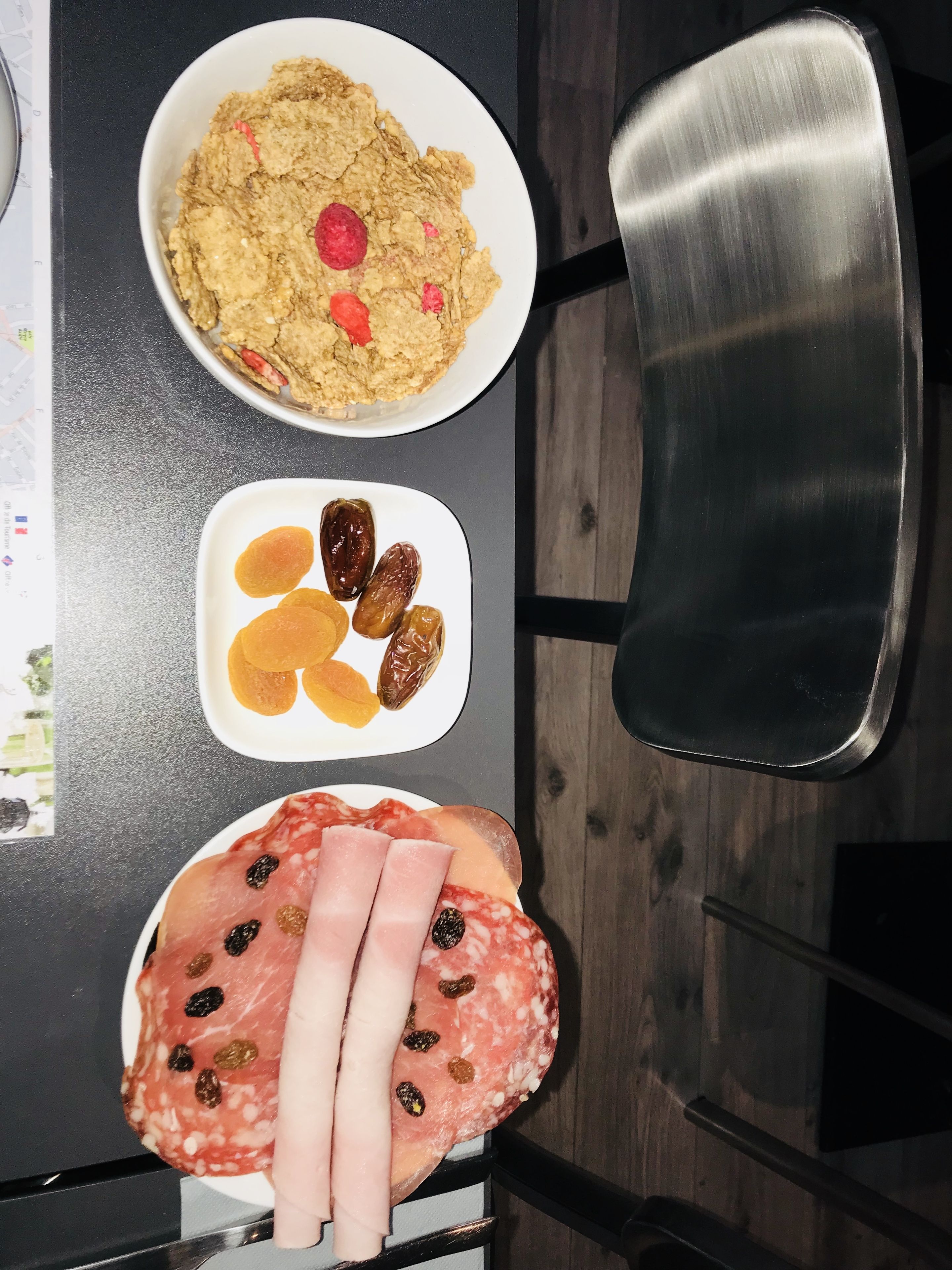 A table with three dishes: a bowl of oatmeal with raisins, a plate of dried apricots, and a plate of salami with olives.