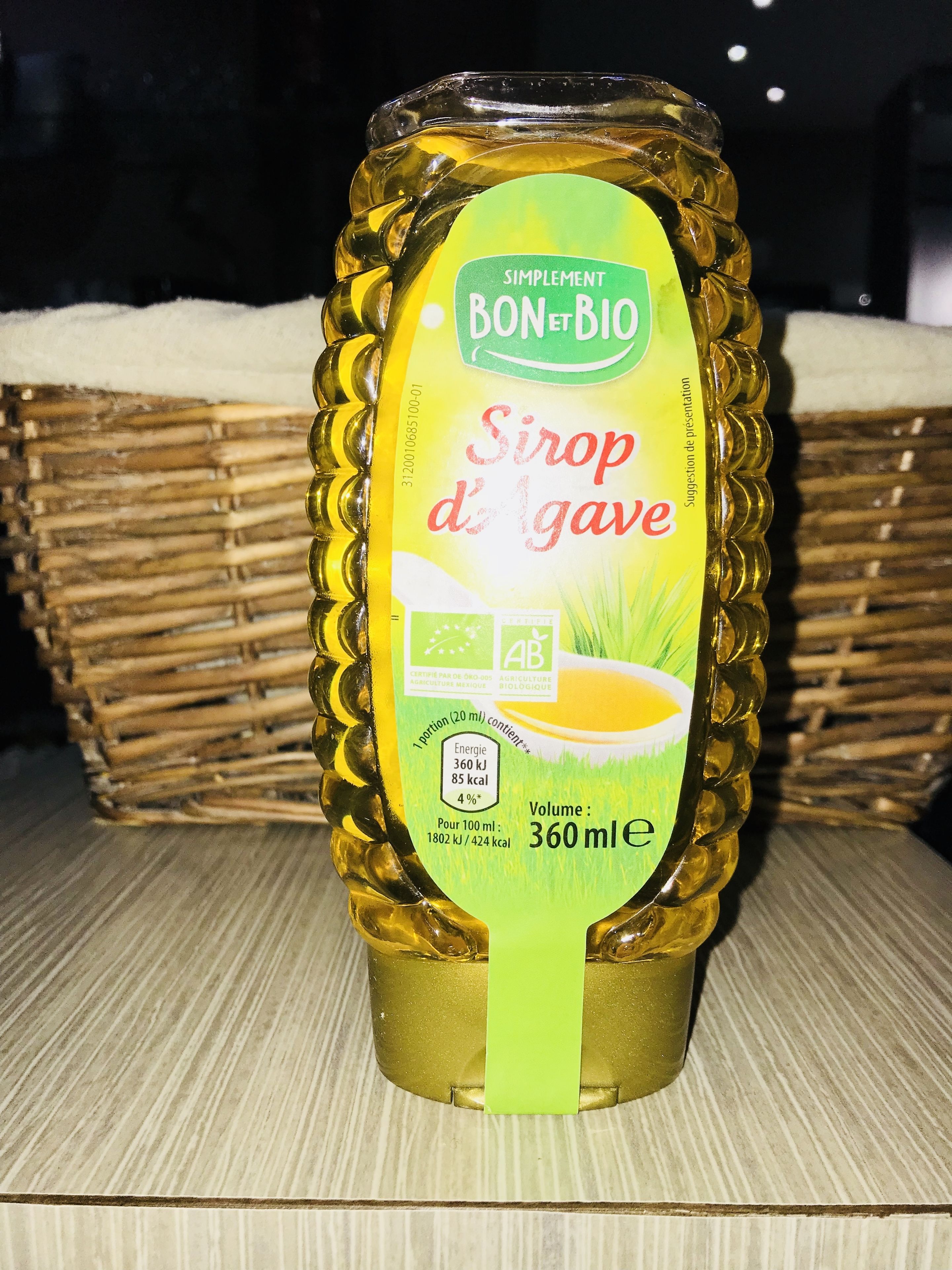 A bottle of Siroppo d'Agave with a green label, placed on a wooden surface with woven baskets in the background.