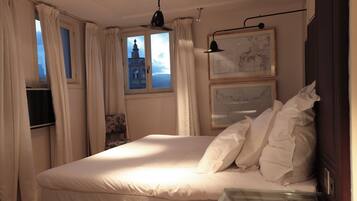 Una cama bien hecha con sábanas blancas, una ventana con cortinas y cuadros enmarcados en la pared.