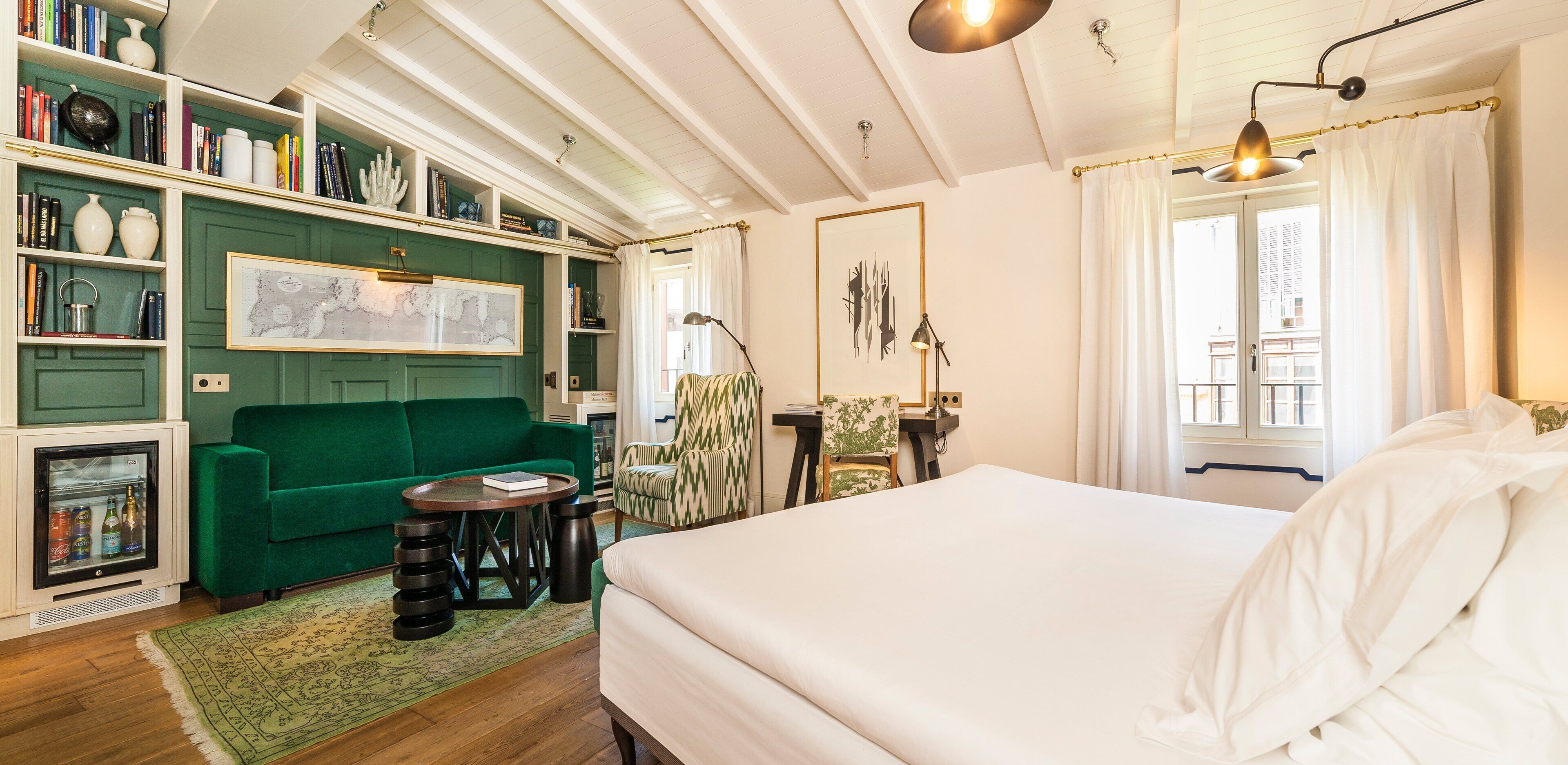 A hotel room with a green sofa, a bed, a small table, and a bookshelf filled with books and decorative items.
