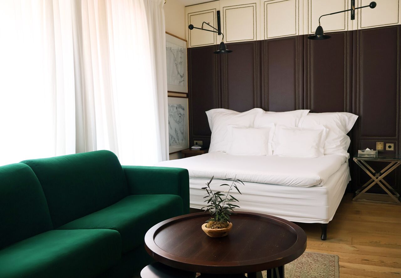 Un dormitorio ordenado con una cama, un sofá verde, una mesa redonda de madera con una planta y una mesita de noche con una lámpara.