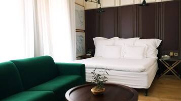 Un dormitorio ordenado con una cama, un sofá verde, una mesa redonda de madera con una planta y una mesita de noche con una lámpara.