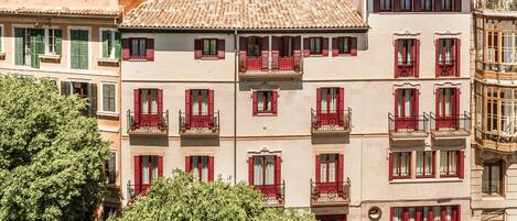 A multi-story building with red shutters and balconies, surrounded by greenery and other buildings.